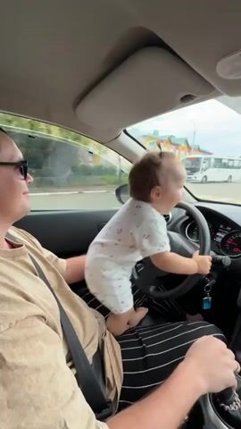 A man wearing sunglasses and a beige t-shirt is in the driver's seat of a car, looking to his left and smiling. A baby wearing a white onesie with red cherries is standing on the man's lap, facing forward and holding onto the steering wheel. The baby is barefoot. The car is in motion. Outside the car window, there are buildings, trees, and a street with traffic. The video shows the interior of the car, the dashboard, and the road ahead. The baby is actively engaged with the steering wheel.