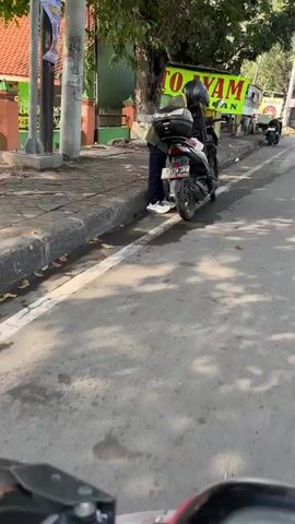 The video shows a person on a black scooter parked on the side of a road. The scooter has a black storage box on the back. The person is wearing a dark jacket, blue jeans, and white sneakers. They are leaning over the scooter, appearing to be adjusting something or preparing to ride. A helmet is on the scooter's handlebars. The road is paved and wet in places. There is a sidewalk next to the road with a curb. In the background, there are buildings, trees, and a sign that says "TO YAM." Another scooter is visible further down the road. The lighting suggests it is daytime.