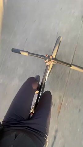 The video captures two individuals cycling on a paved street in Monte Grande, Argentina, during what appears to be either early morning or late afternoon due to the long shadows cast by the bright sunlight.

The first shot is a dynamic, first-person perspective from a bicycle, showing the road with scattered dry leaves. It quickly transitions to a selfie-style view of a young woman riding. She has short, reddish-brown hair and is wearing sunglasses, black earbuds, and black over-ear headphones around her neck. Her outfit consists of a textured black long-sleeved top over a black shirt featuring a white yin-yang symbol, and black leggings. She briefly adjusts her hair and smiles faintly, appearing content. Part of her bicycle frame is visible, showing the word "FIXIE."

The camera then returns to a first-person perspective, looking forward as the woman cycles behind another person. This second individual, seen from behind, is wearing a black shirt, black shorts, and a white, textured backpack. They are cycling down a tree-lined street, with parked cars visible on the left side. The trees have a mix of green and autumnal-colored leaves. The overall atmosphere is peaceful and sunny.
