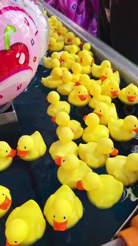 This short video showcases a carnival game, likely a "duck pond" or "rubber ducky race." The primary subjects are numerous bright yellow rubber ducks floating in dark blue water. The ducks have orange beaks and are densely packed, suggesting they are for a game where participants try to pick a winning duck.

In the upper left of the frame, parts of large, colorful balloons are visible, one with a prominent red cherry graphic. To the right, a large, inflatable red and green polka-dotted mushroom or toadstool is partially submerged in the water, also likely part of the game's decor or a prize.

The scene is set at what appears to be an outdoor fair or carnival, with hints of people in the background, visible as legs and feet, suggesting a public event. The lighting suggests daytime, but no specific time or weather conditions can be determined. The overall impression is one of a fun, lighthearted atmosphere typical of a carnival game. There is no visible text in the video.