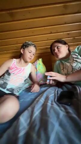 In this media, two individuals, likely a mother and daughter, are positioned on a bed within a room featuring wooden paneled walls, suggesting a rustic or cabin-like setting. The person on the left, a younger individual with braided hair, is wearing a pink and white patterned tank top and black shorts. The person on the right, an adult, is wearing a green t-shirt with a graphic print.

The two are engaged in an activity, with the adult holding a small deck of cards and appearing to interact with them, possibly dealing or showcasing them to the younger person. A smartphone is visible on the bed next to the adult. The lighting suggests it is daytime. The overall mood appears casual and intimate, possibly indicating a moment of shared leisure or a game. The presence of a bed and the interior setting points to a domestic or lodging environment. No visible text is present in the frame.
