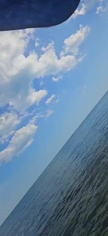 The video pans across a blue sky with scattered white clouds. In the foreground, part of a dark blue umbrella and a person's legs are visible. The ocean stretches out to the right, meeting the sky at a diagonal line. Two metal railings are also visible in the lower portion of the frame, suggesting the perspective might be from a boat or a pier.