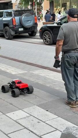 A man wearing a grey t-shirt and blue jeans stands on a sidewalk, looking away from the camera towards the street. He is holding a black object, possibly a phone or controller, in his right hand. In front of him on the sidewalk is a red and black remote-controlled monster truck. Behind him, parked on the street, are two vehicles: a dark grey SUV and a black sedan. The environment appears to be an urban setting with buildings and shops in the background. The sidewalk is paved with rectangular stones, and there is a decorative pattern of linear stones on the street. Several other people are visible in the background, some walking and others standing near the parked cars. The lighting suggests it is daytime.