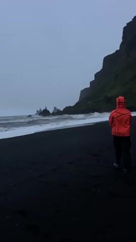 A person wearing an orange hooded jacket and dark pants is walking on a black sand beach. The person is walking away from the camera towards the ocean. The ocean waves are crashing on the shore. In the distance, there are jagged rock formations in the ocean. To the right of the person, there is a tall cliff covered in green vegetation. The sky is overcast and grey.