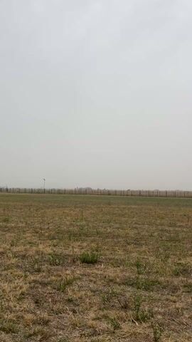 The video shows a large, flat grassy field with a chain-link fence in the distance. The sky is overcast and hazy. There are some small buildings and a light pole visible behind the fence. The grass is mostly brown with some green patches. The camera appears to be stationary. The scene appears to be outdoors.