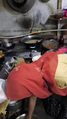 A woman wearing a red hijab and a yellow shirt is bending over a large pot with tongs, stirring its contents. She appears to be in a kitchen with a gas stove and various cooking utensils visible. The stove has a pan with food cooking on it. The kitchen environment looks somewhat cluttered and shows signs of use.