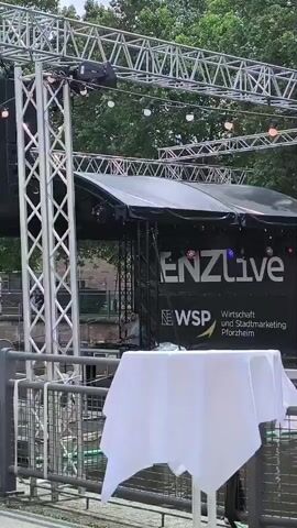 A temporary outdoor stage is set up with a black roof and a large banner displaying "ENZlive" and "WSP Wirtschaft und Stadtmarketing Pforzheim". Several truss structures are visible, supporting speakers and lighting equipment. A white tablecloth covers a round table in the foreground. To the left, several bicycles are parked. In the background, trees and buildings are visible. People are walking along a paved pathway in front of the stage area. One person, a woman, is standing near a large planter with a child. Another group of women are walking towards the stage. The scene appears to be a public event or festival.