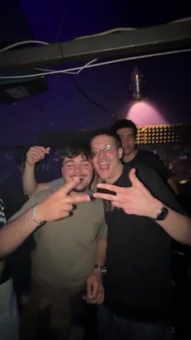 The video shows a group of young men posing in a nightclub with flashing lights.  They are smiling and making peace signs. The camera pulls back to show a DJ booth overlooking a packed dance floor. People are dancing under red and blue neon lights. The walls are decorated with digital displays.