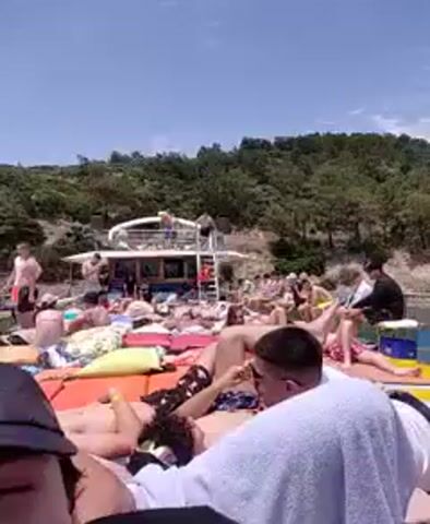 The video captures a lively scene on the deck of a boat, where a crowd of people are sunbathing and socializing under a clear blue sky. A large, white two-story boat is visible in the background, with people standing and sitting on its upper deck, and some using a ladder to descend to the lower deck. The lower deck is packed with individuals, many lying down on colorful mats and cushions, seemingly enjoying the warm weather. The surrounding environment consists of a lush green hillside, indicating a coastal or island setting. The water around the boat appears calm and clear. The overall atmosphere suggests a leisurely day trip or party on the water.