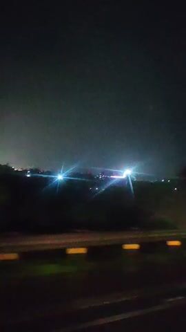 The video shows a nighttime view from a moving vehicle. The scene is dark, with lights from what appears to be a town or city visible in the distance. There are trees and vegetation along the side of the road. The sky is dark and cloudy. Yellow and white markers are visible on the side of the road, indicating the edge of the roadway. The camera is moving, creating a blurred effect in parts of the video.