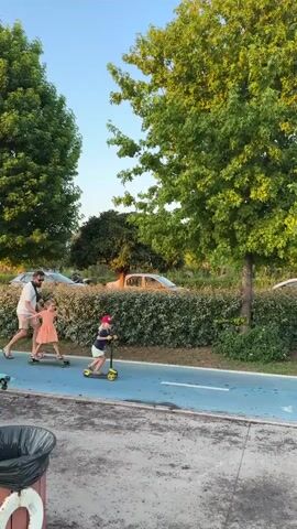 A man wearing a dark shirt and shorts with a beard is skateboarding on a blue path. A young girl in an orange dress is skateboarding next to him. To the right, another young girl wearing a red helmet and shorts is riding a yellow scooter. They are on a paved path with a manicured hedge line behind them. Cars are visible parked behind the hedge. A large tree with green leaves is to the right of the frame. The sky is clear and blue. The scene appears to be outdoors in daylight.