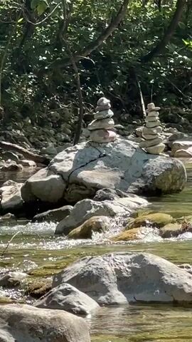 A shallow, rocky river flows gently through a sun-dappled forest. The water is clear and reflects the green of the abundant foliage on the banks. Two cairns, carefully balanced stacks of river stones, stand on larger rocks in the middle of the stream. The left cairn is taller, with more stones, while the right is slightly shorter. The scene is tranquil and natural, with no people or animals visible.