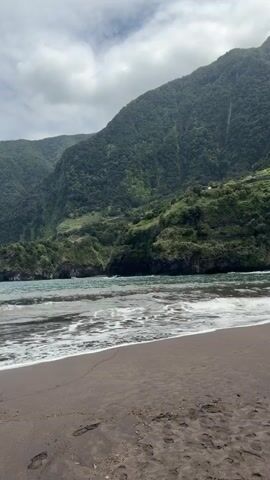 The video shows a beach with dark sand, ocean waves rolling in, and a large, green mountain in the background. The sky is cloudy. There are footprints visible on the sand. The water is a mix of blue and green. The mountain is covered with dense vegetation.