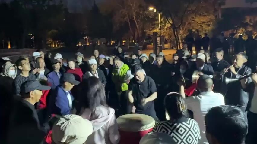 At night in a park, a crowd of around 40 adults are watching and listening to music. Many of the people are wearing hats and casual clothing. In the center of the crowd, a man in a black shirt is drumming on a large, red-rimmed drum. To the right of the drummer, a man in a dark shirt is playing a wind instrument, possibly a Suona. Other people seem to be clapping and moving to the music. The park is dimly lit by streetlights, and the background is blurred with trees and foliage. Some people in the back of the crowd seem to be standing still and observing.