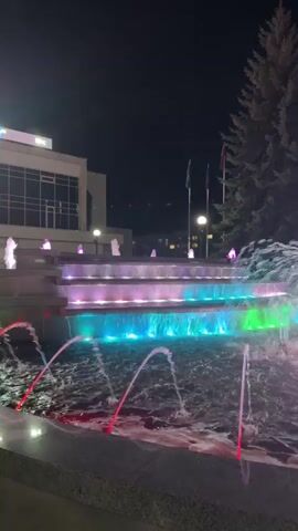 The video shows a colorful fountain at night. The fountain has multiple tiers and is illuminated by vibrant LED lights in shades of pink, purple, green, and blue. Water streams from various jets, creating a dynamic display. In the background, a building with large windows and flags can be seen, along with a tall evergreen tree. The overall scene is one of a well-lit urban environment at night, focusing on the illuminated fountain.