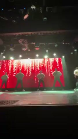 A low-angle shot depicts five dancers from the waist up on a stage bathed in red and green light. Their backs face the camera, obscuring their faces. Each dancer wears a tan sweater and black pants, standing with legs spread and hands resting on their hips. Behind them, a large screen displays a red backdrop with golden sparks. The stage, illuminated by stage lights, has visible cracks. Above, more stage lights and a disco ball hang. An indistinct voice says "Players" and "Watch out, Take it, Ready".