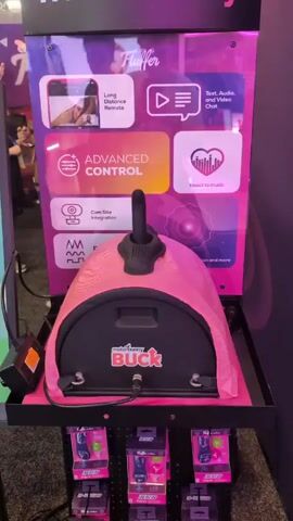 The video shows a booth at a trade show for the adult toy company "Fluffer." The booth features various sex toys, including a pink, arched device with a joystick on top called "Buck," as well as a table displaying several differently-shaped insertable sex toys. Multiple screens are set up showing promotional material and presumably demonstrating the products. There are several people at the booth, including booth workers and attendees. One woman is wearing bunny ears and holding one of the products. There are multiple other attendees visible in the background walking through the trade show. The booth design is primarily black and pink, with promotional banners and displays highlighting features like "Long Distance Remote" and "Advanced Control."