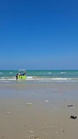 The video shows a person on a beach flying a kite. The beach is sandy with some debris visible. The ocean is in the background with waves and the sky is clear and blue. There are a few fishing boats in the distance. The person is standing and holding the string of the kite, which is white and shaped like a bird. The kite is flying in the wind. There is a red and white bucket next to the person.