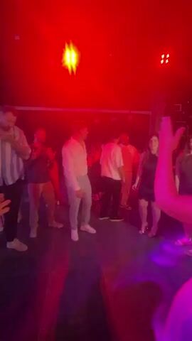 A group of people are dancing in a dimly lit club or venue. The lighting is primarily purple and blue. In the foreground, a person's hands are visible, suggesting they are holding a phone or camera to record the scene. In the middle ground, several individuals are dancing. A man in a white shirt and light-colored pants stands out, seemingly observing or directing. To his left, a man in a striped shirt and another in a dark t-shirt are also present. To the right of the man in white, a woman in a black dress is dancing, followed by another woman in a dark dress. In the background, more people are visible, along with the stage lighting and the dark ceiling of the venue. The atmosphere appears to be a social gathering or party.