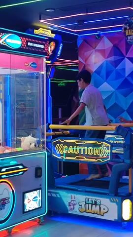 The video shows an indoor arcade with neon lights and arcade machines. In the center of the frame, a young boy wearing a light-colored shirt and jeans is standing on a platform with yellow handlebars, appearing to be playing a game. To his right, a woman in a dark top and jeans is standing and looking at the boy. Behind the boy, there's a wall with a geometric, colorful pattern. To the boy's left, a large claw machine with a white teddy bear inside is visible. The floor is decorated with cartoon characters and text that says "LET'S JUMP." The overall environment is dimly lit, with the bright neon lights of the arcade machines creating a vibrant atmosphere.