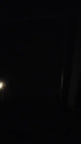 This video is shot in low light conditions, likely at night. A faint yellow light source is visible on the left side of the frame, possibly a street light or car headlight. The scene appears to be outdoors, with the presence of what looks like a dark, indistinct landscape or ground in the lower portion of the frame. On the right side, a vertical element, likely a wall or a structure, is partially illuminated by a thin streak of light. The upper portion of the frame is dark.