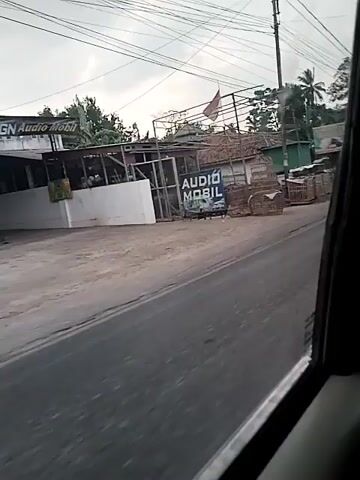 The video shows a blurry street view from inside a vehicle. On the right side of the street, there are several buildings and signs. One sign reads "AUDIO MOBIL" with a picture of a car stereo. Another larger sign appears to be an archway with text that is partially obscured. There are also trees and utility poles with many wires. A truck is seen driving past on the left side of the road, causing motion blur. The sky is overcast.