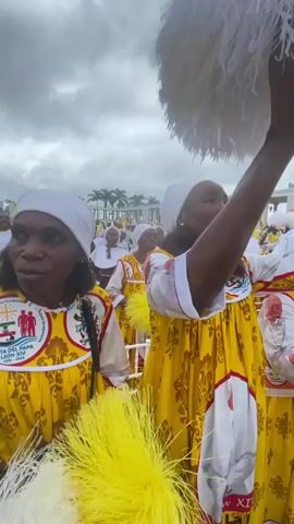 Several women are seen attending an outdoor gathering under an overcast sky. They are uniformly dressed in long yellow dresses with white sleeves and yokes, adorned with a repeating pattern. Each dress features prominent circular patches; one on the left shows a graphic with the text "VISITA DEL PAPA JUAN XIV 2023," while others display a portrait of a Pope, resembling John Paul II, on both the chest and sleeve areas. The women also wear white headscarves. One woman in the foreground holds a bright yellow feathery pom-pom, and another holds a white one. In the background, palm trees and large, white architectural structures are visible, suggesting a public square or event venue. The scene appears to be a religious or commemorative event related to a papal visit.