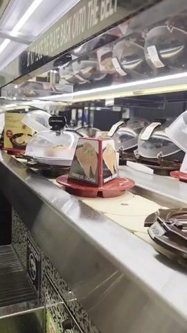 The video shows a close-up, eye-level shot of a sushi conveyor belt in a restaurant. The belt is moving, carrying plates of sushi. In the foreground, there is a metallic counter. Several sushi plates are visible on the belt, some covered with clear plastic domes. One plate has a card in front of it with an image of salmon and the text "UMAMI OIL SALMON." Another plate has a card with an image of ramen and text that includes "MAZEMEN." Above the belt, there is a black sign with white text, partially obscured, that reads "BACK ONTO THE BELT". The lighting is bright, likely from overhead fluorescent lights. There are no people visible in the frame. The overall scene suggests a typical experience at a kaiten-zushi (conveyor belt sushi) restaurant.