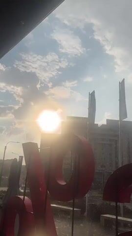 The video shows a close-up of a large red letter "V" in the foreground, with a sunny sky and clouds in the background. The sun is visible, creating a lens flare. In the middle ground, there are buildings, flags on poles, and some trees. The foreground is wet, with what appears to be rain or water falling. The video pans slightly to the right, revealing more of the red letter and the environment behind it. There are several people visible in the middle ground, some standing and some seated. Cars are also present, driving on a road. A green structure, possibly a small building or booth, is visible. The overall scene suggests an outdoor environment, possibly a plaza or public space, during a time when it is raining.
