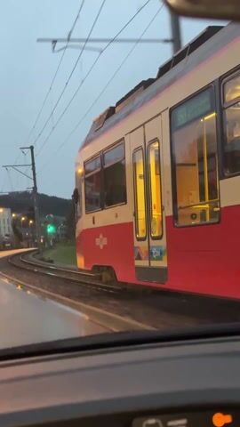 A vehicle's dashboard view captures a red and white train making a turn on tracks alongside a wet road. Streetlights and a green traffic light illuminate the scene, with buildings and trees visible in the background under a grey sky. The train's destination, "Stallikon," is displayed in the windows.