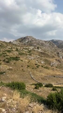 This is a landscape video showing a mountainous region with dry grass and scattered bushes. In the distance, there are rugged mountains under a partly cloudy sky. There are no people or visible animals in the video. The video appears to be a panorama or a slow panning shot of the natural scenery. There are some stone walls visible on the slopes, suggesting past human activity or land management. A few small structures, possibly cabins or sheds, can be seen in the mid-ground of the landscape.