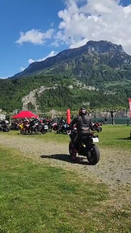 The video shows a group of motorcycles parked on a grassy area near a lake. There are people standing around and near the motorcycles, some getting ready to ride. The area is near a body of water. In the background there are mountains with some snow on top. The sky is blue with a few white clouds. A red canopy is visible in the left side of the video.