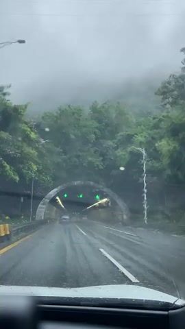 The video shows a view from inside a car driving through a tunnel on a rainy day. The car's windshield is wet, with water droplets visible. The tunnel is illuminated with green and white lights, and there are orange traffic cones visible along the side of the road. Trees and fog are visible outside the tunnel entrance. Another car can be seen ahead in the tunnel.