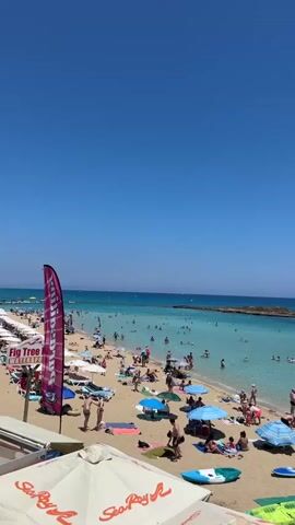 The video shows a sunny day at a crowded beach. People are swimming in the clear turquoise water, relaxing on the sandy beach under umbrellas, and engaging in water sports like paddleboarding and jet skiing. A pier extends into the water with several boats docked. The sky is a vibrant blue with no clouds. The scene depicts a typical summer vacation atmosphere with a focus on leisure activities.