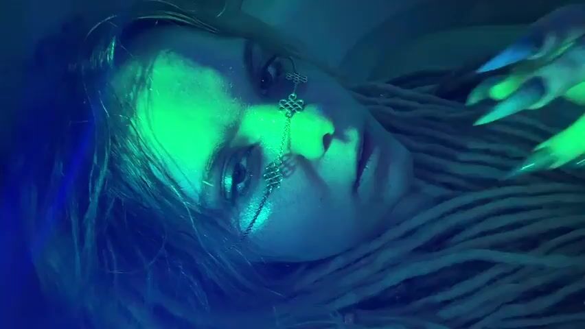 The video is a close-up, low-angle shot of a young woman lying down, her head tilted to the side. She has long, light-colored dreadlocks. Her face is partially illuminated by green light, casting dramatic shadows. A delicate silver chain with knot-like designs is draped across her face, connecting her cheekbones. Her eyes are open, and she appears to be looking towards the camera or slightly off to the side. One of her hands, with long, pointed fingernails, is raised near her face, with her index finger touching her lips. The lighting is dark and atmospheric, with prominent blue and green hues, creating a somewhat mystical or artistic effect. The background is indistinct and appears to be dark or out of focus.