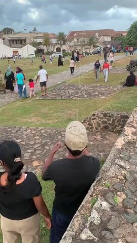 The video shows an outdoor scene with several people in the foreground and middle ground, and buildings and sky in the background. Two individuals, one wearing a black shirt and a baseball cap, and another wearing a black shirt and a ponytail, are in the immediate foreground. They appear to be looking up at something in the sky. In the middle ground, a grassy area is visible with paved pathways. Several people are walking and standing on the grass and pathways. Some individuals are dressed in casual clothing, while others, particularly women, are wearing headscarves and modest attire. In the far distance, there are buildings with red tile roofs, suggesting a town or historical area. The sky is partly cloudy with a mix of blue sky and grey clouds. The video focuses on the people in the foreground and their apparent observation of something in the sky. A kite can be seen flying high in the sky. The overall atmosphere appears to be relaxed, with people enjoying an outdoor activity.