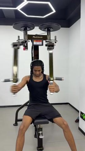 A young man is exercising at the gym. He is sitting on a machine, holding on to two handles. He is moving them back and forth in front of him. He is wearing a black tank top, black shorts, and headphones. The gym is well-lit and has white walls. There are weights on top of the machine.