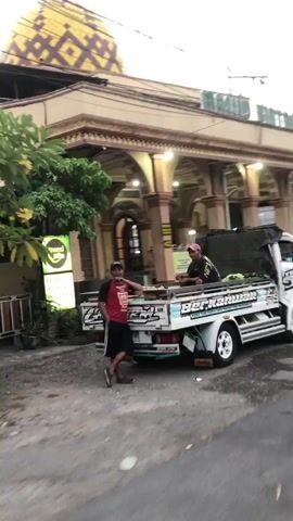 Two men stand beside a white flatbed truck with the word "BERKAH" written on its side. The man on the left wears a red shirt with white lettering and black shorts. He is leaning against the truck. The man on the right wears a black shirt with white and pink lettering and a pink hat. He is holding a cell phone. The truck has green lights underneath its bed. In the background, there is a building with an arched entryway and a domed roof decorated with yellow and white patterns. There are trees and a neon sign to the left of the building. The ground is paved.