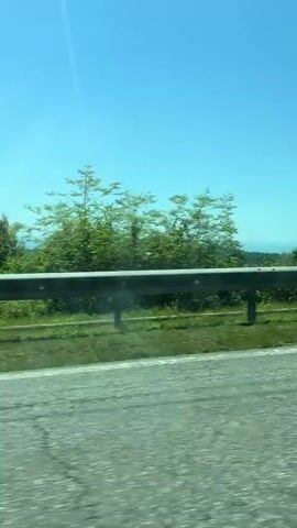 A daytime video shot from a moving vehicle captures a scenic landscape passing by. The primary focus is the roadside, featuring a gray metal guardrail running horizontally across the lower middle of the frame. Beyond the guardrail, lush green grass, bushes, and trees dominate the view. In the distance, undulating green hills or mountains can be seen under a bright, clear blue sky. The sun is visible as a bright glare in the upper left corner, indicating a sunny day. The road surface, which appears to be asphalt, is visible in the foreground, showing movement. The setting is identified as Viadati Eshyra, Abkhazia. There is no visible text or identifiable people in the video.