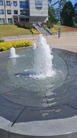 The video captures a serene outdoor scene featuring an active water fountain in a plaza. The fountain, centrally located, sprays a powerful jet of water upwards, creating a dynamic splash. In the background, a modern, multi-story building stands tall, with a sloping green lawn leading up to its entrance. Yellow-flowering bushes and green shrubs line the edge of the plaza and the lawn, adding vibrant color to the landscape.

Several flags are visible on the building, including the Latvian flag. A crest is also present on the building's facade. In the distance, a street with a pedestrian crossing and traffic signs can be seen, suggesting an urban or semi-urban environment. The sun appears to be shining, indicating a clear and pleasant day. The overall mood is tranquil and refreshing, with the sound of the splashing water creating a calming ambiance. The focus is on the natural beauty of the water and its interaction with the surrounding architecture and landscaping. No people are visible in the frame. The time of day is likely daytime, given the bright lighting. The location cues point towards Ogre, Latvia.