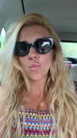 A woman with long blonde hair and sunglasses is in the passenger seat of a car. She makes a kissy face and then smiles at the camera. The video then shows the view from the front windshield of the car, which is driving down a road surrounded by trees and greenery. The sky is cloudy.