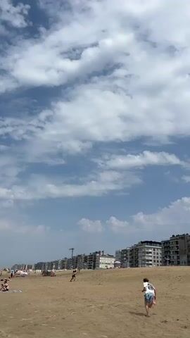 The video shows a wide shot of a sandy beach with a line of apartment buildings in the background. The sky is blue with scattered white clouds. Two children are playing on the beach in the foreground. One child is running away from the camera, wearing a white tank top and blue shorts. The other child, further in the background, appears to be running towards the camera. The buildings in the background are multi-story structures with balconies. The scene is a bright, outdoor day at the beach.
