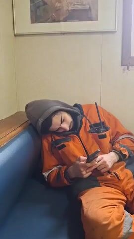 The video shows a man in orange work overalls relaxing in a room. Initially, he is lying on a blue couch with a grey hooded sweatshirt pulled over his head, looking at his phone. The camera pans to the right, revealing a small desk with papers and various items on it. The man sits up, still looking at his phone, and settles into a chair at the desk, legs crossed. The room has a window, a small cabinet, and various papers and items attached to the walls.