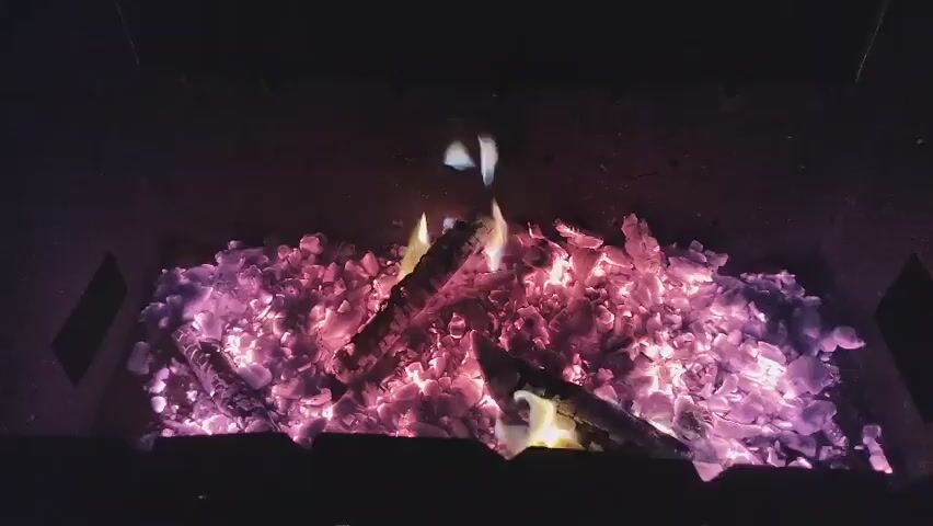 The video shows a close-up view of a fire pit filled with glowing embers and burning wood. The embers are predominantly pink and purple, with some brighter red and orange spots. Several pieces of wood are positioned on top of the embers, with visible flames and smoke emanating from them. The background is dark, suggesting the footage was captured in low light conditions. The camera appears to be positioned directly above the fire pit, looking down. The scene is static, with no discernible movement other than the flickering of the flames and the shifting of the embers.