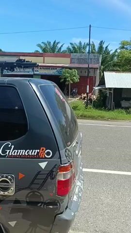 The video begins with a close-up of the rear of a dark-colored car, showing a "Glamour PRO" sticker and some graphic designs. The car is parked on the side of a road. The camera then pans to the right, revealing a rustic, dilapidated building with a damaged roof and open sides, situated next to the road. Beyond this structure, there is lush green vegetation and trees. The camera continues to pan and tilt, showing a winding asphalt road in the foreground, bordered by green grass and dirt. In the distance, a motorcycle with a rider is seen traveling on the road. The background features a range of majestic, green mountains under a bright blue sky with scattered white clouds. The scene suggests a rural or semi-rural environment, likely in a tropical or subtropical region. The overall atmosphere is peaceful and natural, with signs of human presence in the form of the road, the car, the buildings, and the motorcycle.