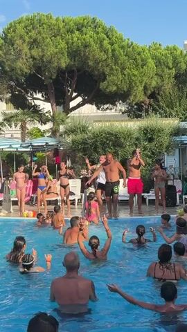 A group of people are gathered around a swimming pool. Some are in the pool, while others are standing on the deck. There are children and adults, many of whom are wearing swimwear. Some people in the pool are splashing and playing. On the deck, a few individuals appear to be leading an activity, possibly a dance or game, as they are gesturing and moving energetically. One person in the background is wearing a white shirt and black shorts, and another is in red shorts. The environment appears to be outdoors, with trees and some buildings visible in the background. The lighting suggests it is daytime, likely sunny.