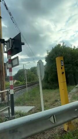 The video shows a view from inside a car, looking out at a railroad track and the surrounding environment. The car is moving. The railroad tracks are on the left side of the screen, with overhead power lines. There is a signal light with red and white stripes. In the distance, there are trees and houses. A "Danger" sign is visible on the right side of the screen. The interior of the car shows a dashboard with a phone mounted on a holder. The weather appears to be cloudy.
