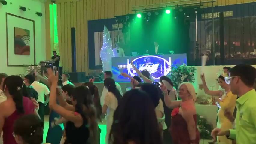 The video captures a lively wedding reception held indoors in Vera, Spain. A diverse group of people, dressed in festive attire, are enthusiastically dancing on a dance floor bathed in vibrant green and blue lighting. The atmosphere is energetic and celebratory, with many attendees raising their arms and enjoying the music.

In the background, a DJ booth is set up with a large, illuminated screen displaying the text "JOSU BALTA DARE". A DJ is actively working the turntables, contributing to the party's ambiance. Above the dance floor, the ceiling is draped with white fabric, creating a soft, ambient glow, and string lights add a touch of sparkle.

The room features a stage area with more lighting equipment and a large screen or backdrop. Artwork is visible on the walls, suggesting a decorated venue. The time of day appears to be evening or night, given the indoor setting and artificial lighting. The overall mood is one of joy and celebration, typical of a wedding reception.