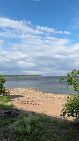 This is a vertical, eye-level shot of a scenic waterscape on a cloudy day. The foreground is a textured beach of pebbles and sand, with some scattered rocks. Lush green foliage frames the right side of the image. Beyond the beach, a wide, calm body of water stretches towards a distant, tree-lined shore.

Several buoys, marked with pink and white caps, are visible in the water, suggesting a marked course or navigational area. A small, dark boat with what appears to be one or two occupants is moving across the water. In the far distance, faint white shapes suggest other boats or structures.

The sky is a bright blue with scattered, fluffy white clouds, indicating fair weather with some cloud cover. The overall impression is one of a serene and pleasant summer day at the water's edge. The presence of the distant shore and buoys implies a large lake or bay, possibly near a settlement, given the context of Shlisselburg, Russia, which is known for its fortress on an island in Lake Ladoga. The lighting suggests it is daytime, likely late morning or afternoon. There is no visible text in the media file.