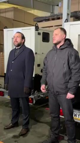 Two men stand in a warehouse beside two enclosed generators on trailers. The man on the left wears a long blue coat, dark pants, gloves and brown shoes. He has a beard. The man on the right wears a dark colored jacket, dark colored jeans and dark boots. He speaks and gestures with his right hand. The generators are white. Pallets of wood are visible in the background.